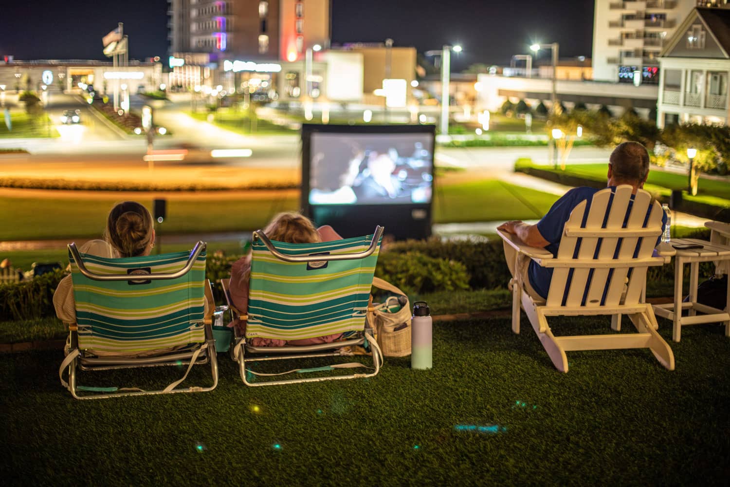 A group of people sitting in lawn chairs watching a movie on a big screen outdoors