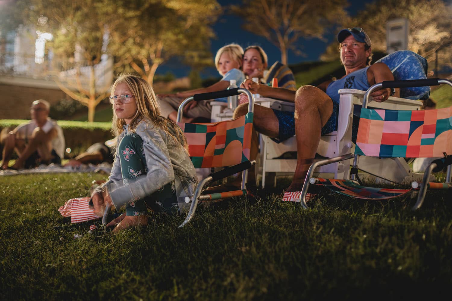 A family sitting on a lawn watching an outdoor movie at night time